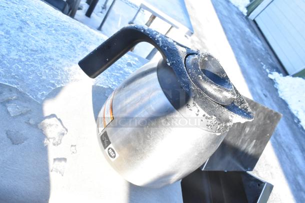 Stainless steel trash can shell with a swinging flap lid, shown in snowy outdoor setting, NSF label visible.