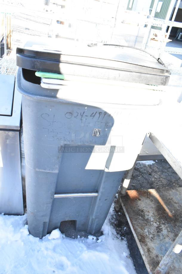 Poly trash can containing multiple cutting boards, visible on top. Exterior shows writing and minor wear, set outdoors.