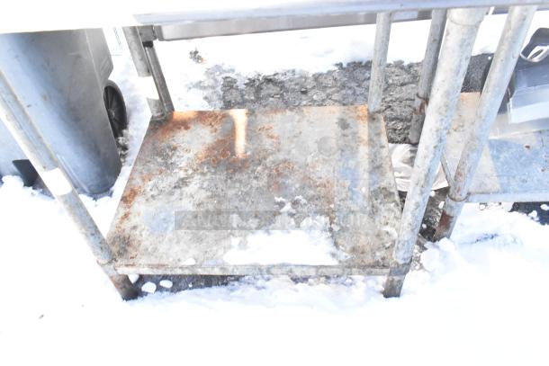 Stainless steel table with metal under shelf, showing signs of rust and wear, set outdoors on snowy ground.