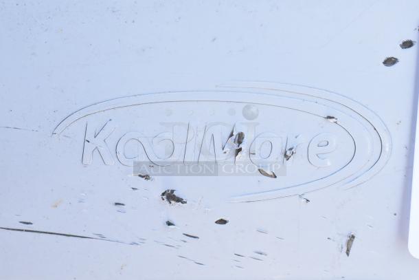 Engraved "KoolMore" logo on stainless steel surface, showing scratches and dents on a commercial sink panel.