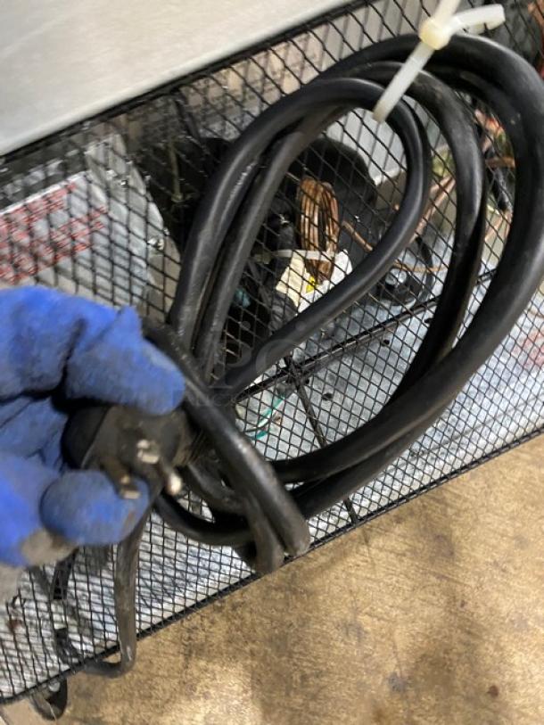 Coiled black power cord with a plug, held in a gloved hand, against a wire mesh background.