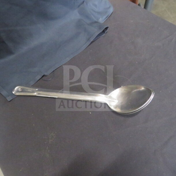 Stainless steel spoon, model 3XBID, with a polished finish on a dark fabric surface. Slightly used condition.