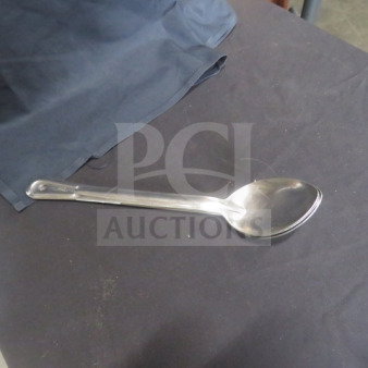 Stainless steel spoon, model 3XBID, with a polished finish on a dark fabric surface. Slightly used condition.