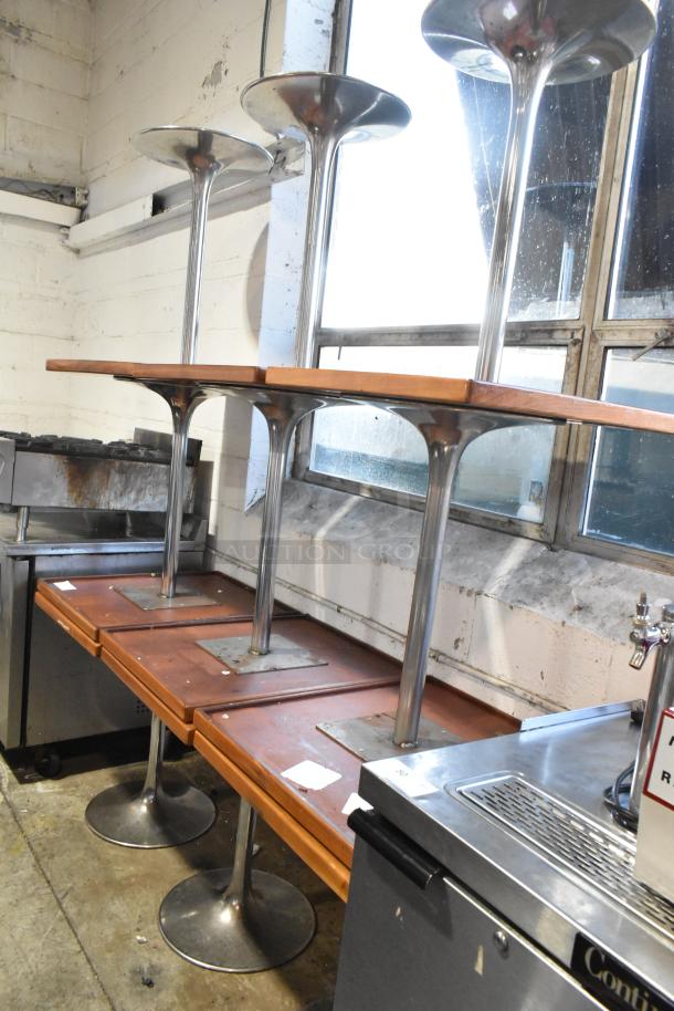 9 dining height tables, wooden tops, chrome finish bases, stacked in storage. Visible wear, suitable for restaurants.