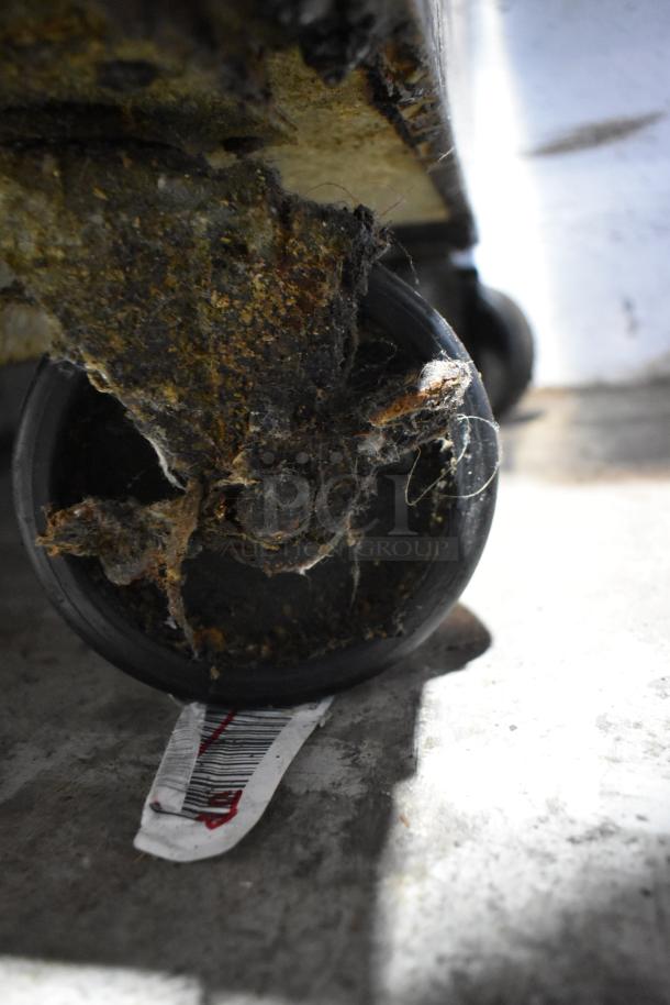 Close-up of a wheel under a True TWT-27F freezer, showing dust and debris buildup. Barcode tag visible. Condition needs cleaning.