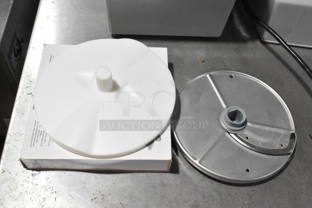 Robot Coupe R2N processor parts on metal surface; includes a white plastic lid and a metal slicing blade attachment.