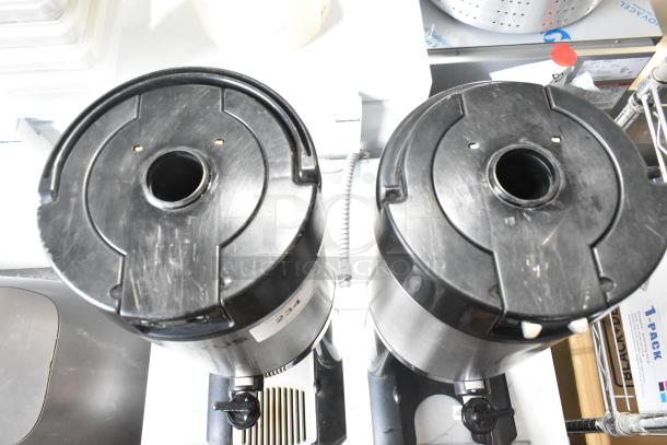 Two metal countertop beverage holder dispensers with black lids, in used condition, featuring dispensing spouts and visible labels.