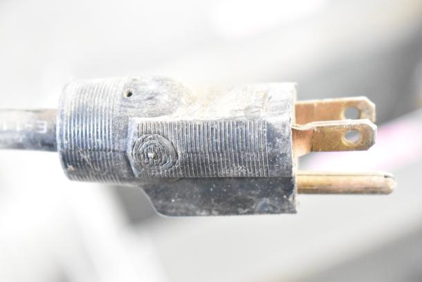 Close-up of a worn electrical plug, showing two parallel prongs and a grounding pin. It is connected to a device operating at 115 volts, 1 phase.