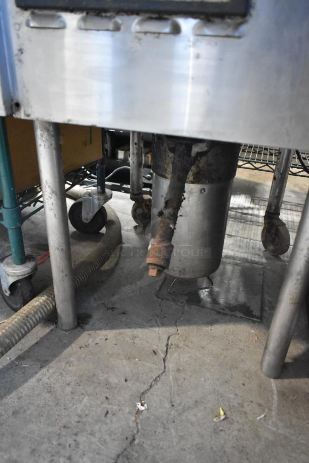 Stainless steel commercial pressure deep fat fryer with visible wear and rust. Features robust metal legs and drainage hose.