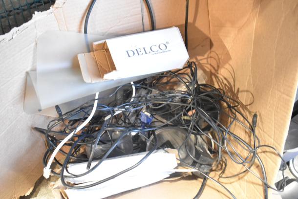 Lot of miscellaneous electronics in a box, featuring cables and a Delco box. Includes cameras, speakers, and phone accessories.