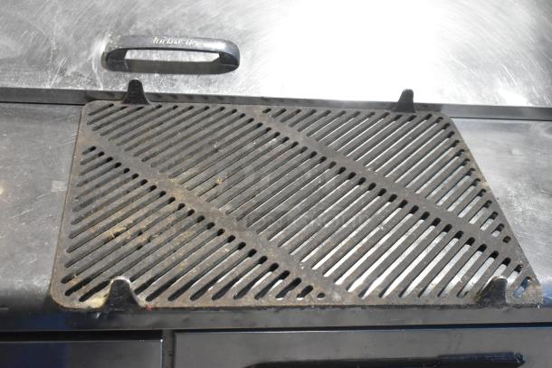 Metal grate, rectangular, slightly worn, diagonal slats, dark finish, four corner supports, suitable for industrial use.