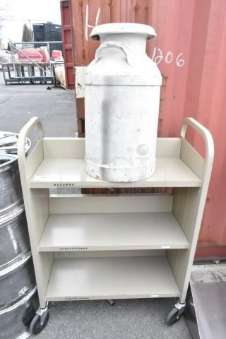 Tan metal 3-tier cart on commercial casters, featuring a large metal jug. Labels on shelves read "Returns" and "Acquisitions."