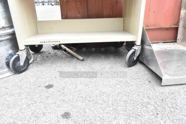 Tan metal 3-tier cart on commercial casters with metal jug, shows scuff marks. Marked "ACQUISITIONS" on lower shelf.