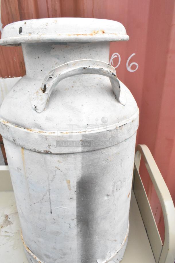 Tan metal 3 tier cart with a large, weathered metal jug on commercial casters. Signs of rust and wear visible.
