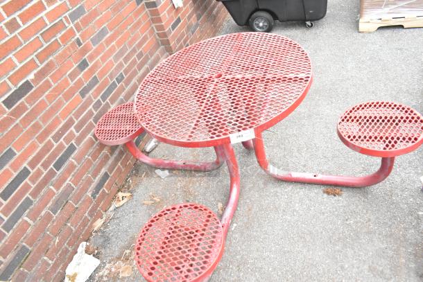Red round mesh table with attached three seats, weathered condition, located outdoors near a brick wall.