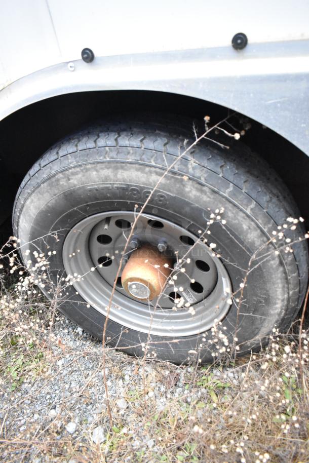 Close-up of a tire on a Stuff4less 24' fast food trailer, showing tread and rim, surrounded by dry grass.