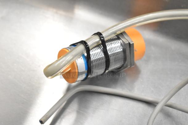 Metallic sensor or connector component with orange and blue accents, secured with zip ties, possibly part of cookie machinery.