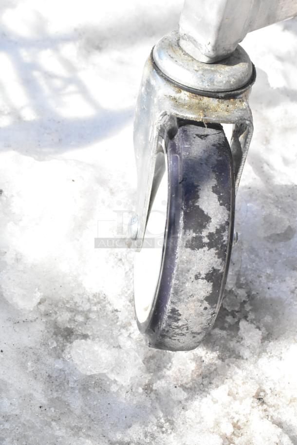 Close-up of a commercial caster wheel attached to a pan transport rack, showing signs of wear on a snowy surface.