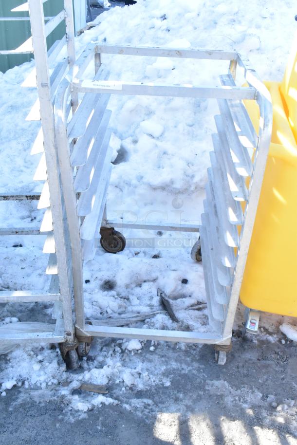 Commercial pan transport rack with metal shelves and commercial casters. Snowy outdoor setting. Markings show “PCAuctions.com.”