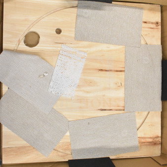 Three boxes containing a wooden board with cutouts, covered with textured fabric patches, shown in a cardboard box.