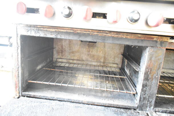 American Range commercial gas-powered 6 burner stove with flat top griddle, 2 ovens, back splash, and over shelf. Visible signs of use.