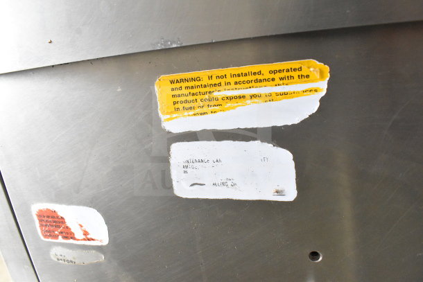 Commercial floor-style manual tilt braising pan with visible wear. Labels with safety warnings and maintenance care instructions.