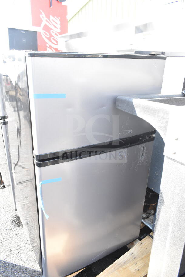 Silver mini cooler, possibly Galanz brand, shows light wear with visible tape and scratch marks. 115 volts, 1 phase.