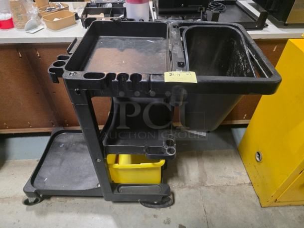 Black janitorial cart with three shelves, attached waste bin, and a yellow bucket. Used condition, showing slight wear.