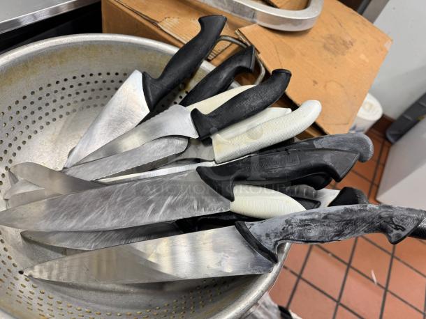 A collection of knives with black and white handles in a metal colander. Some handles have visible brand markings "Wüsthof." Knives are in used condition.