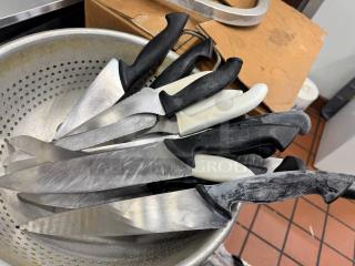 A collection of knives with black and white handles in a metal colander. Some handles have visible brand markings "Wüsthof." Knives are in used condition.