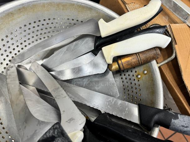 Multiple kitchen knives with various plastic and wooden handles, placed in a metal colander. Knives show signs of use.