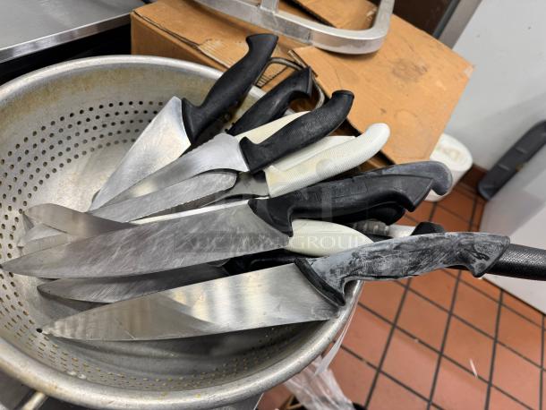 Assorted kitchen knives with black and white handles in a metal colander. Some knives show wear and patina. Various brands, including Wüsthof.