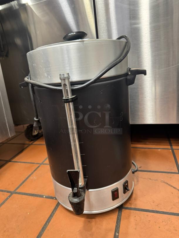 Stainless steel commercial coffee urn with a black finish, visible power switch, and spigot, showing signs of use.
