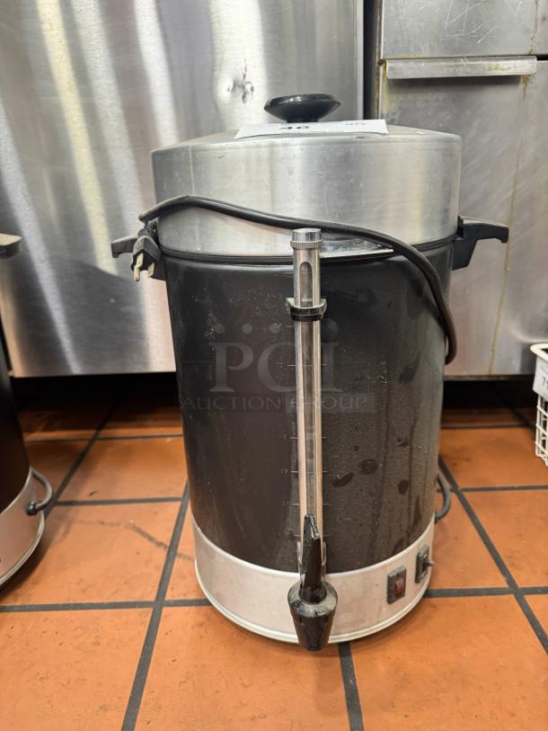 Large black and silver coffee urn with a side spigot, power cord, and indicator light, showing signs of wear.