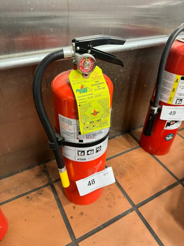 Red fire extinguisher with a pressure gauge, yellow inspection tag, and instructional label. Good condition, labeled Pacific Fire.