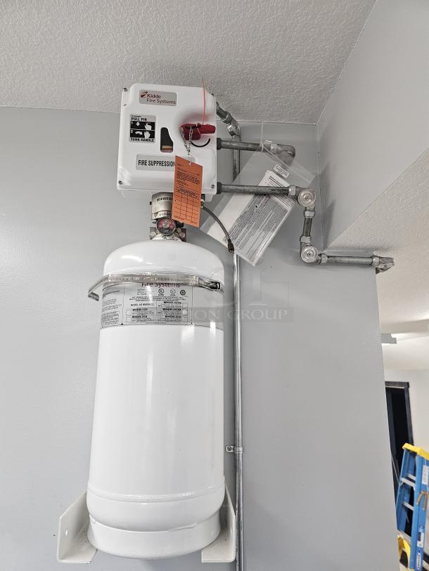 Kidde fire suppression system with white tank and control box, featuring safety labels and manufacturer tag, mounted on a wall.