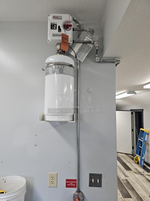 Kidde fire suppression system, mounted on wall, white cylinder tank, manual release label, visible inspection tags.