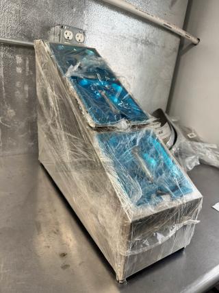 Brand new stainless steel countertop condiment dispenser, wrapped in protective plastic, on a metal surface.