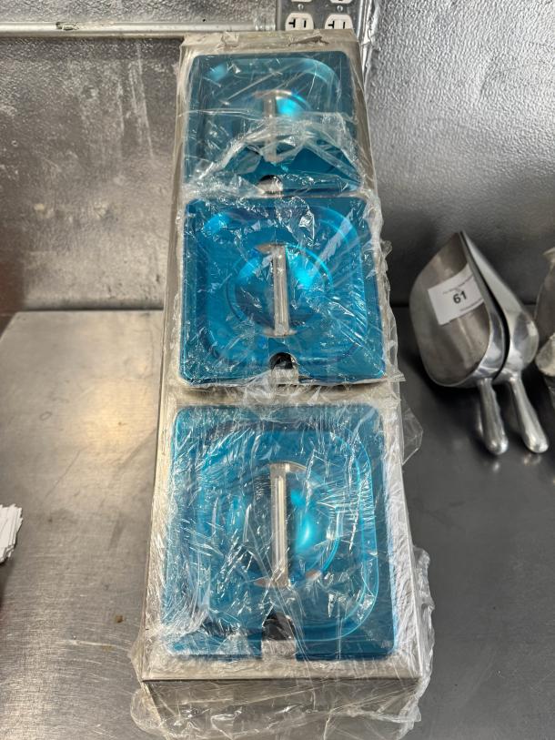 Stainless steel countertop condiment dispenser, brand new, with protective blue film and wrapped in plastic. Three compartments.