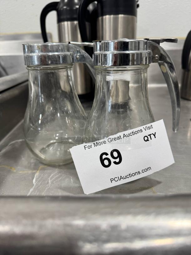 Two glass syrup dispensers with chrome lids, shown slightly used. Auction tag reads "69" from PCI Auctions.