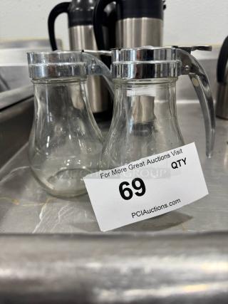 Two glass syrup dispensers with chrome lids, shown slightly used. Auction tag reads "69" from PCI Auctions.