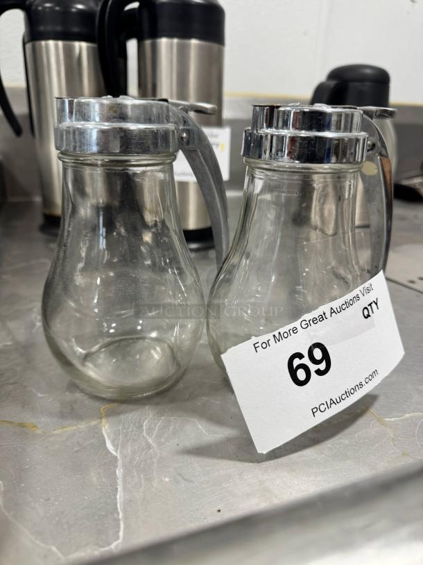 Lot of 2 glass syrup dispensers with metal lids, good condition, includes auction tag "69" from PCI Auctions.