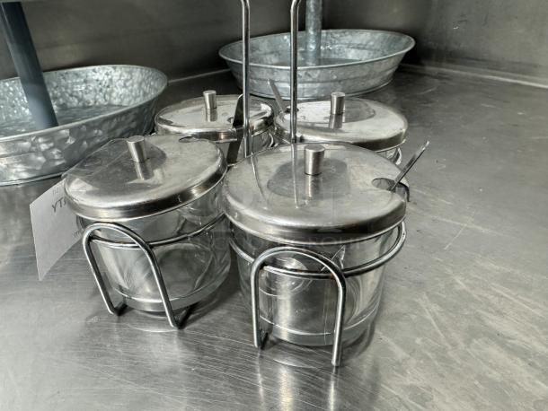 Glass and metal condiment caddy with four jars, lids, and spoons. Good condition, ideal for kitchen or dining use.
