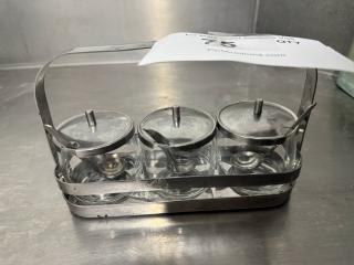 Glass condiment caddy with three jars, metal lids, and spoons in a metal holder; appears in good condition.