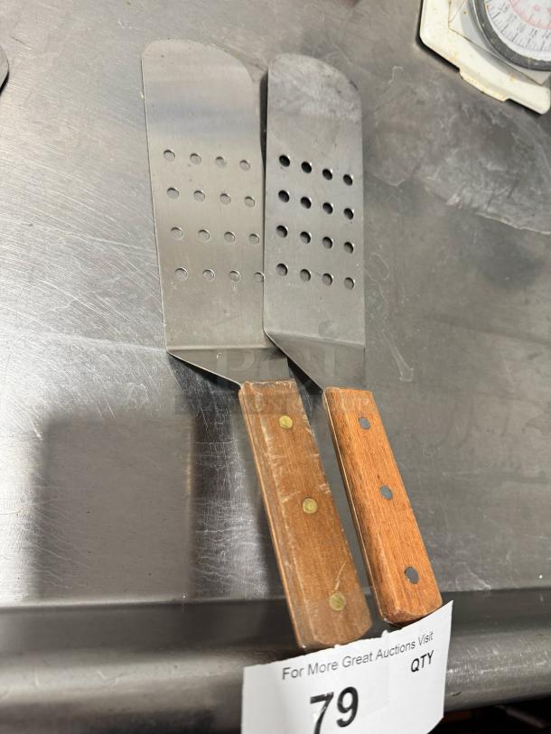 Two perforated turners with wood handles, metal heads with round holes, slight wear visible, tag marked "79."