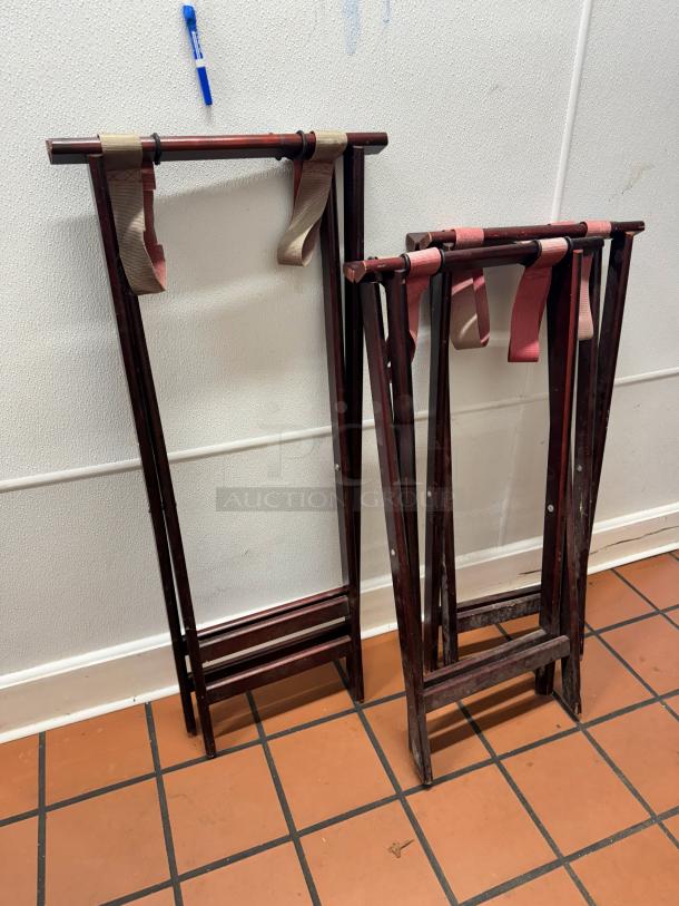 Two folding wood tray stands with brown straps, standing against a white wall on a tile floor.