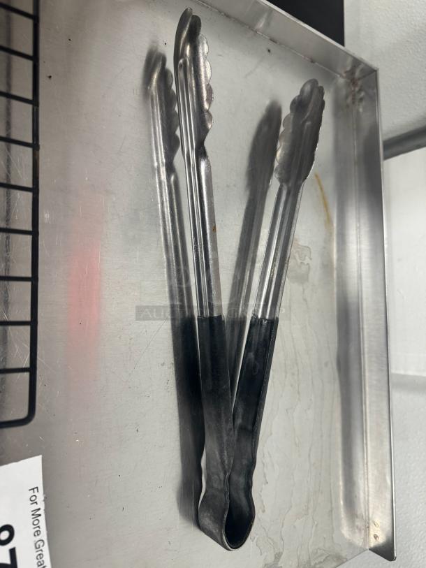 Metal kitchen tongs with black grip handles, used condition. Suitable for various cooking tasks. Item: TONGS BIDX5.