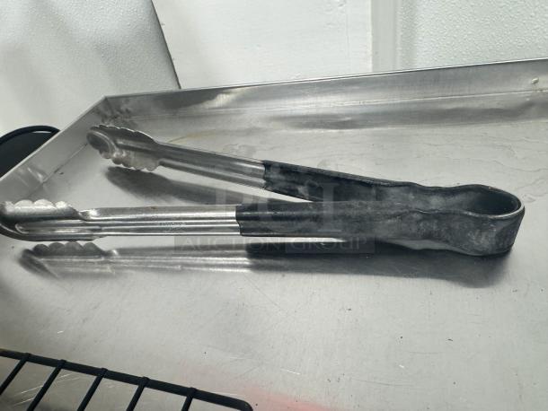 Stainless steel tongs with black rubber grips, model BIDX5, in good condition on a metal surface.