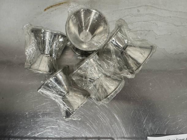 Lot of 5 stainless steel double jiggers, wrapped in clear plastic. Appears unused, suitable for measuring cocktails.