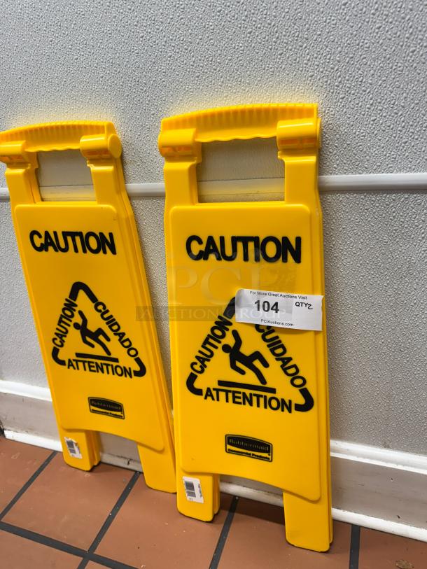 Yellow Rubbermaid floor sign with "Caution" in English, Spanish, and French. Tag reads "104, PCIauctions.com."